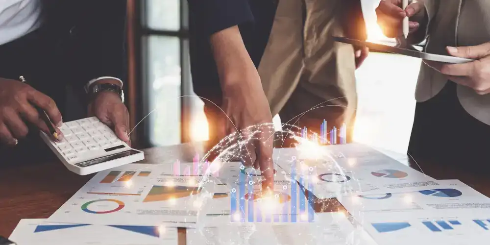 Profissionais de negócios analisando dados financeiros em uma mesa com documentos impressos contendo gráficos e estatísticas. Uma pessoa usa uma calculadora enquanto outra aponta para os relatórios. Projeções holográficas de gráficos e visualizações de dados emergem dos documentos, criando um efeito de realidade aumentada que simboliza análise de dados moderna e tomada de decisões baseada em informações.